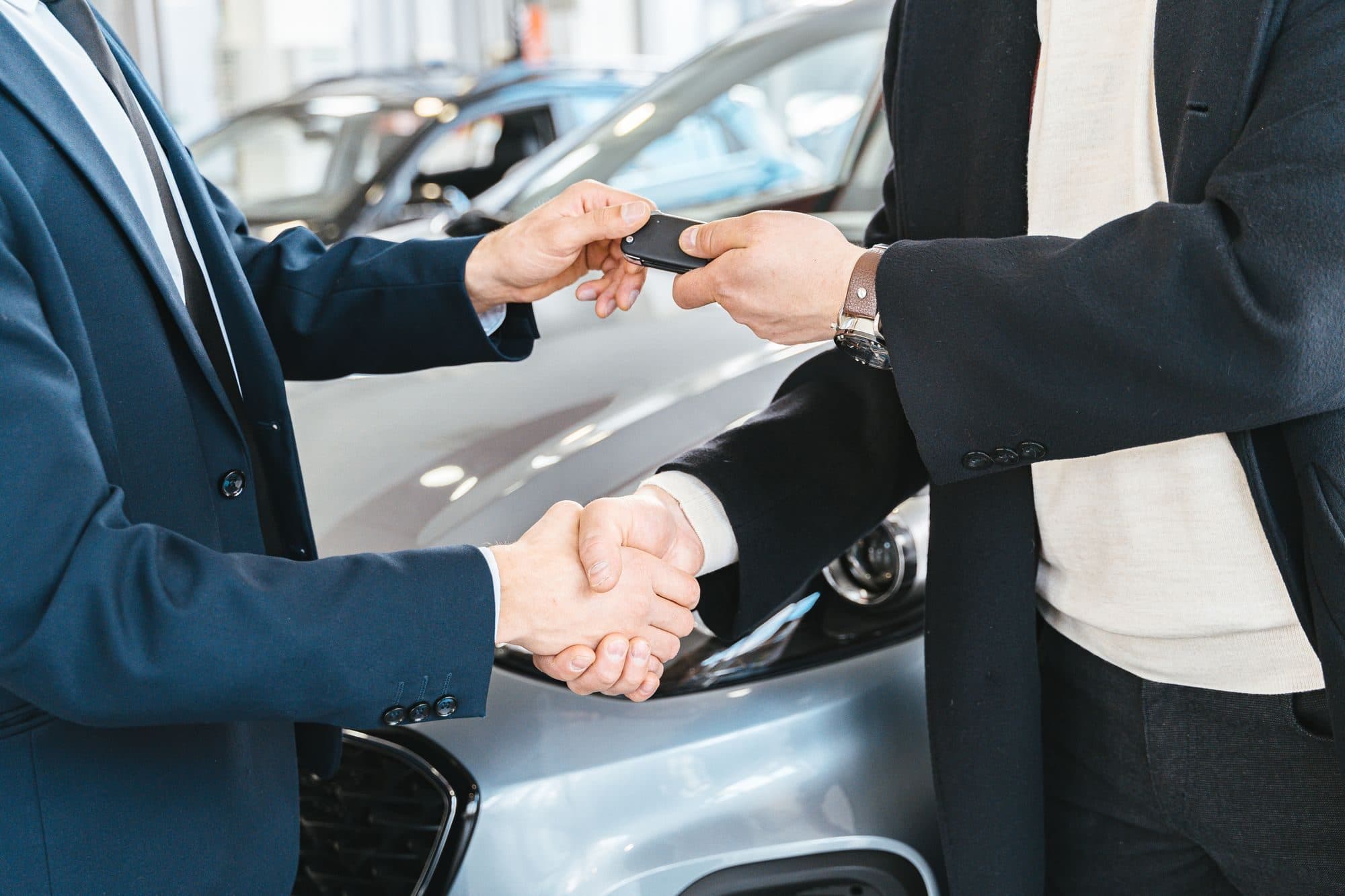 Aperto de mãos e entrega de chave de carro entre duas pessoas de terno diante de um veículo, cena de compra financiada com gravame