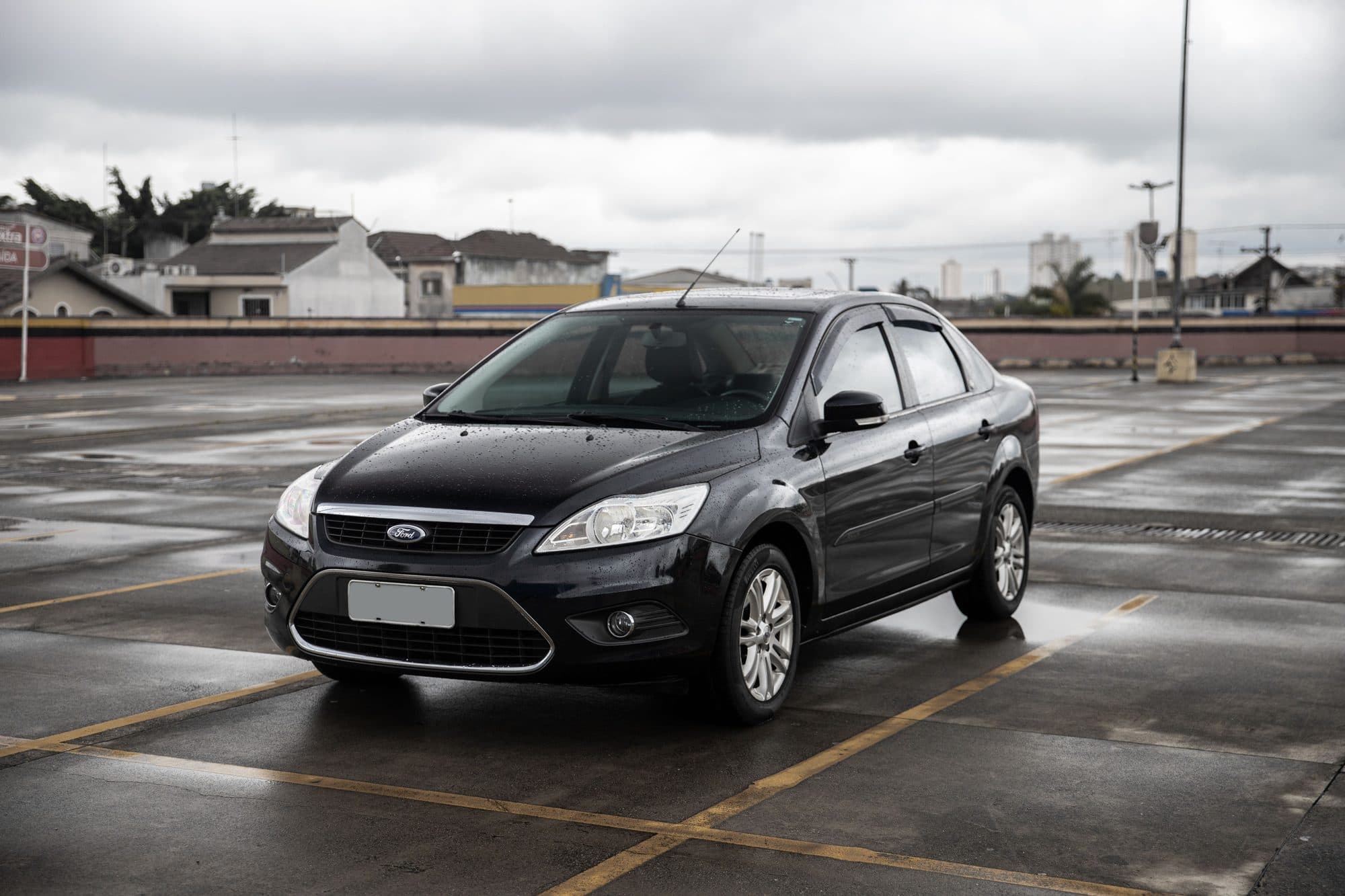 Quanto vale o seu carro: sedã preto usado estacionado em um pátio molhado de chuva, visto de frente, com a placa sem caracteres legíveis